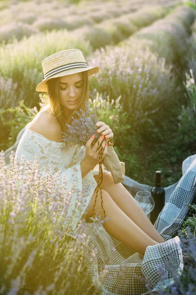 A girl in a dress and hat sits on a lavender field in the sun — 图库照片