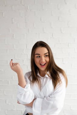 Şaşırmış güzel esmer, beyaz bir duvarın arka planında elini kaldırıyor.