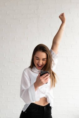 Kız telefona bakar ve sevinçle bağırır. Elleri havada, beyaz arka planda.