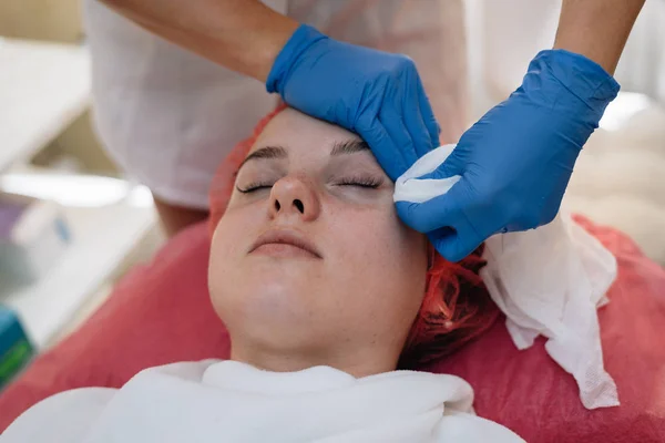 Estetista pulisce il viso della donna con un tovagliolo prima della procedura, gli occhi della paziente chiusi, primo piano — Foto Stock