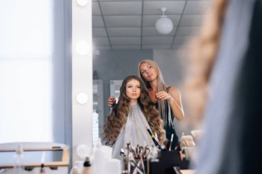 Makyöz, uzun saçlı modelin saç stilini düzeltir. Kızlar aynadaki yansımalarına bakar.