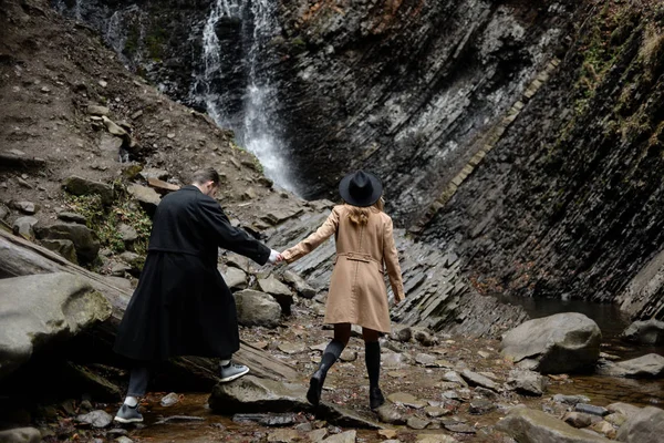Couple élégant homme et femme portant un manteau et chapeaux marchent dans la nature près d'une cascade. Voyage de noces en Islande — Photo