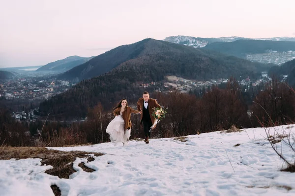 一对新婚夫妇在雪地里跑来跑去 男人的外套是棕色的 花束是为婚礼准备的 度蜜月的人在滑雪胜地度假 放在你的文字位置上 — 图库照片