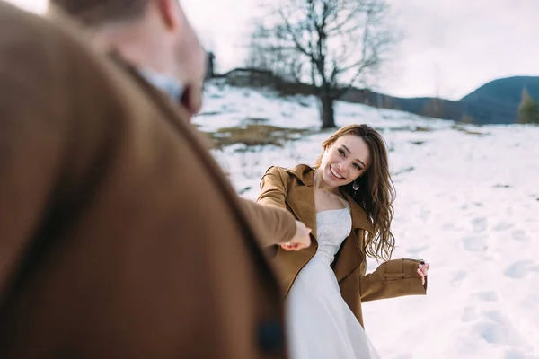 Flirty bride holds the hand of her man. They are dancing in the snow in the forest. — Stockfoto