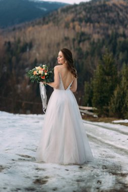 cute bride in a white dress with a neckline on her back holds a bouquet on a background of a winter forest.