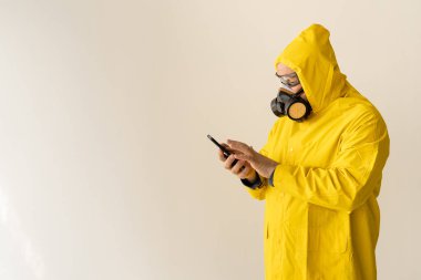 A man in a yellow chemical protection suit and a face mask clicks on the phone. Dialing a number. Quarantine isolation concept. Alone at home. place for text clipart