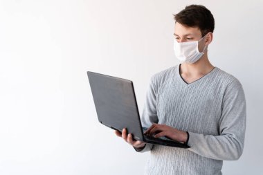 Maskeli genç adam elinde dizüstü bilgisayarla evde dikiliyor. Yüzüne koruyucu bir tıbbi maske takılmış. Portret evde. Coronavirus kontrol konsepti. Uzak Karantina covid-19