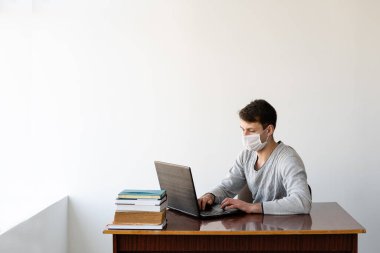 Tıbbi maskeli genç adam dizüstü bilgisayarda çalışıyor, evdeki masasında oturuyor. Salgın ve karantina sırasındaki eğitim kavramı. Online eğitim. Metin için yer