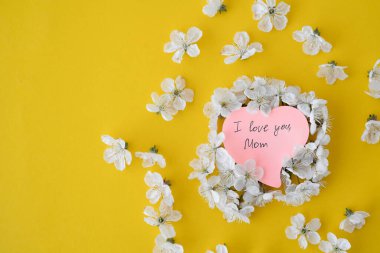 Bir kiraz ağacının çiçeği beyaz arka planda, kalp şeklinde pembe bir yaprağın üzerinde yatar. Seni seviyorum anne. Metin için yer