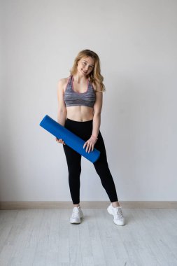 Fitness yoga egzersizi yapan kadın. Ellerinde yoga minderiyle mutlu bir kadın parlak bir odada beyaz bir duvara karşı duruyor. metin için yer