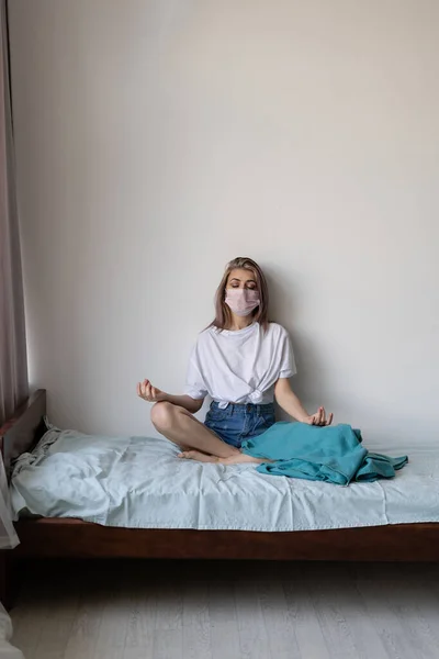 Rest at home during quarantine. Young beautiful woman is sitting on the bed. Meditation at home. Vertical frame. place for text. Face mask