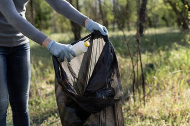 Lastik eldiven takan kadın eli plastik bir şişeyi çöp torbasına atıyor. Gönüllü, çöpü temizler. Çevre kirliliği. Bölgeyi temizlemek için.