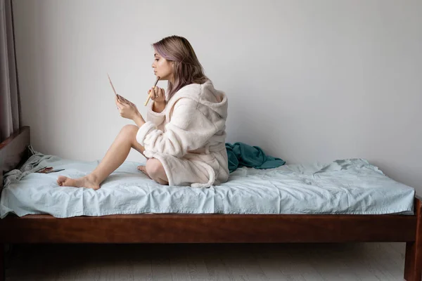 Joven hermosa mujer se sienta en un albornoz en la cama y se pone maquillaje a sí misma. Muro blanco lugar para el texto . —  Fotos de Stock