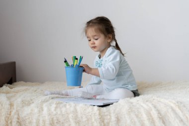 Avrupa görünümlü küçük bir kız evde yatakta oturuyor. Kırtasiyeli bir sepet aldı, parlak keçeli kalemlerle çizmek istiyor..