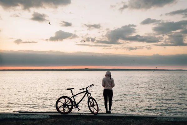 A young sporty girl stands on the seashore and looks into the distance, she rests from a long bike ride, next to a sports bike, thinks about herself and her life, sports and health for her is life.
