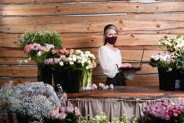 entrepreneur during a pandemic and quarantine. Woman florist in a protective mask on her face stands near a table with flowers. It works remotely. Accepts an order for bouquets online.