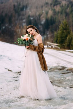 Bir buket çiçekli şık genç gelin güzel bir günün tadını çıkarıyor. Gelin kışın, temiz havada, dağların arasında duruyor..