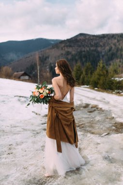 Güzel gelin sırt üstü çıplak omuzlarıyla duruyor ve elinde bir buket düğün çiçeği tutuyor. Çekici gelin, bir kış ormanının arka planında çekiliyor..