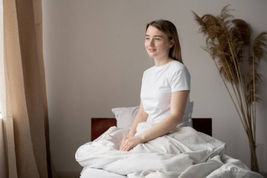 Genç ve güzel bir kadın yatakta yalnız oturur ve güzel bir günün hayalini kurar. İşinizi sabah erkenden planlayın. Yüzünde gizemli bir gülümseme.