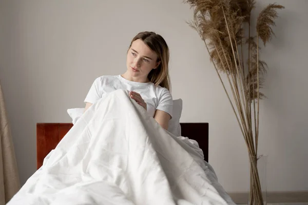 Romantic girl with a sly smile in a white T-shirt sitting on the bed. Portrait of a dreamy cute young woman relaxing in bedroom in the morning. White wall, place for text.