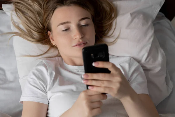 Closeup portrait of a young woman in bed. Mobile phone in the hands. Read news before bedtime online via smartphone