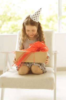Pencerenin yanında oturan ve elinde kırmızı kurdeleli bir kutu tutan mutlu kız. Karantina doğum günü. Tatil sırasında bir çocuk.
