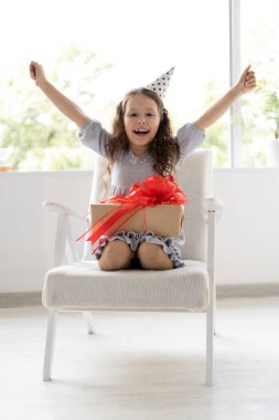 . Şenlik şapkası kafaya giydirilmiş. Çocuğun yüzünde neşe ve neşe var. Sevgili kızın için güzel bir hediye. Büyük bir hediye kutusu, kırmızı güzel bir fiyonk.