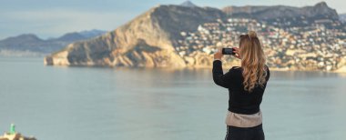 Akıllı telefon tutan kadın, dağların yamacındaki Calpe kasabasının ve Akdeniz 'in fotoğraflarını çekiyor. Anı yakala, turistin yeni yerleri ziyaret edişinin tadını çıkar
