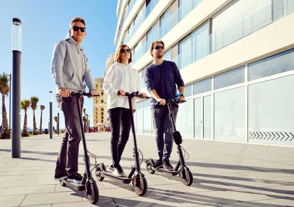 Three best friends young 20s -30s girl and guys spend time outdoors gathered together driving on electric scooter