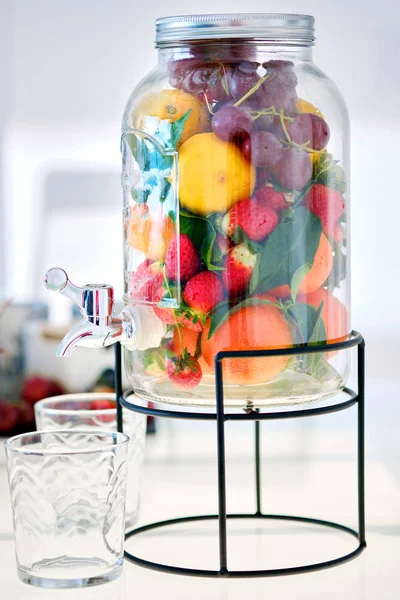 Close up view glass mason gallon jar beverage dispenser with stand filled with fresh ripe multi coloured yummy fruits and berries and two empty glasses, no people. Healthy beverages easy preparation