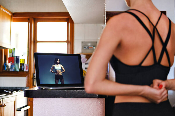 Rearview woman working out at domestic home kitchen using computer on-line training, pc screen view coach showing exercises trainee repeat them. Exercises sport from home concept