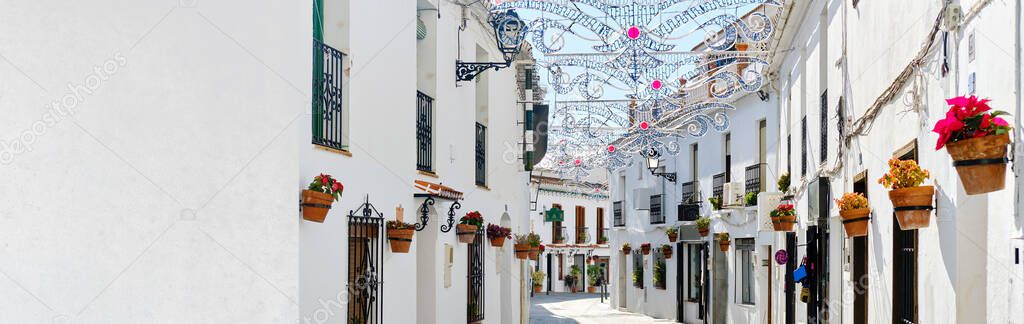 Panoramic image white copy space view, empty street famous village of Mijas in Spain. Charming narrow streets with New Year decorations, hanging flower pots on walls, no people. Costa del Sol, Malaga