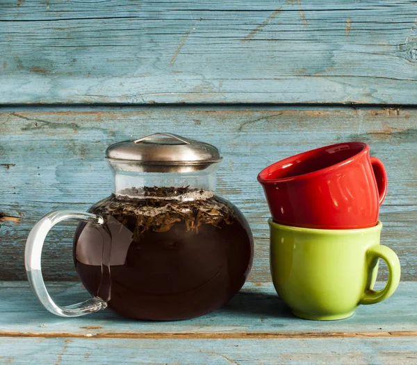 Průhledná skleněná čajník s černého čaje a barevné šálky na dřevěný stůl — Stock fotografie