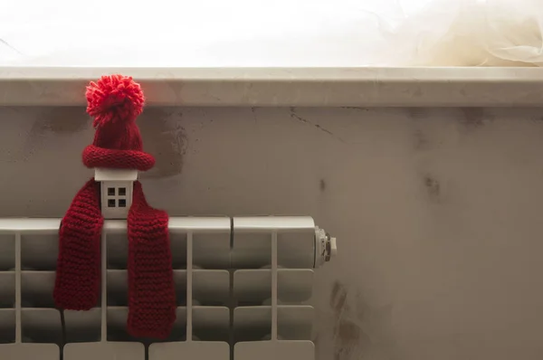 暖气散热器上戴着红色针织帽子和围巾的房子模型. — 图库照片