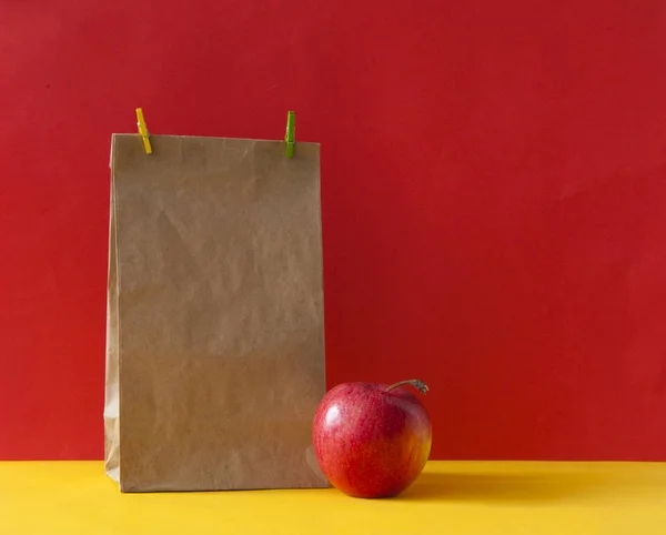 Saco de papel marrom e uma maçã vermelha no fundo de papel colorido — Fotografia de Stock