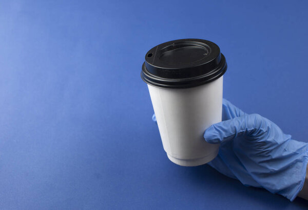 female hand in blue medical glove holding paper coffee cup on blue background with copypsace 