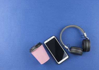 Kulaklıkların üst görüntüsü, akıllı telefon ve mavi arka planda kağıt bardak kahve. Kopyalama alanı olan kulaklıkların minimalist fotoğrafı.