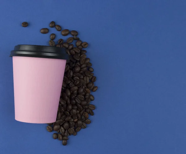 Pink paper coffee cup with a plastic black lid on a blue background with coffee beans. Coffee cup with take-away coffee. Copyspace, flat lay.