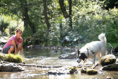 Genç bir kadın ormandaki sığ nehir suyunda köpeğiyle oynuyor..