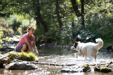 Genç bir kadın ormandaki sığ nehir suyunda köpeğiyle oynuyor..