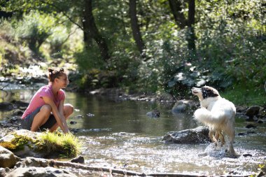Genç bir kadın ormandaki sığ nehir suyunda köpeğiyle oynuyor..