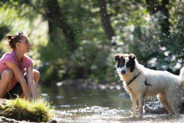 Genç bir kadın ormandaki sığ nehir suyunda köpeğiyle oynuyor..