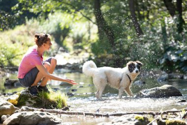 Genç bir kadın ormandaki sığ nehir suyunda köpeğiyle oynuyor..