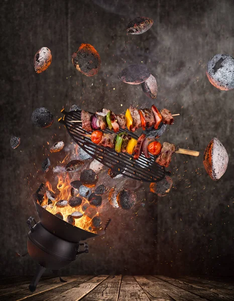 Grelha de chaleira com briquetes quentes, grelha de ferro fundido e espetos saborosos voando no ar . — Fotografia de Stock