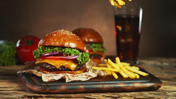 Frytki spadają obok cheeseburgera, leżącego na drewnianej desce do krojenia. Super wolny ruch — Wideo stockowe