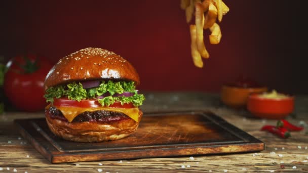 Franse frietjes vallen naast cheeseburger, liggend op vintage houten snijplank. Superlangzame beweging — Stockvideo