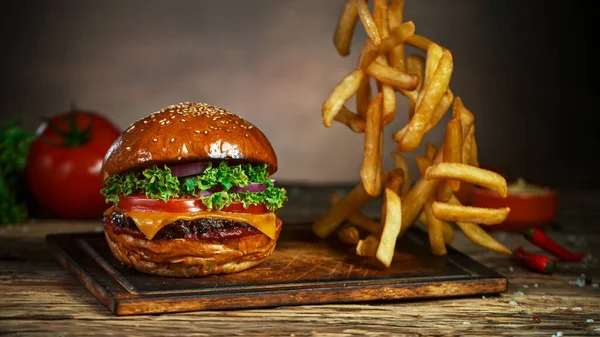 Batatas fritas caem ao lado de cheeseburger, deitado na placa de corte de madeira vintage. — Fotografia de Stock