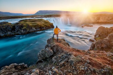 Adam gün batımında İzlanda 'da inanılmaz Godafoss şelalesinin yanında duruyor.