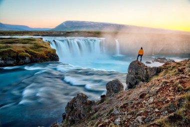 Adam gün batımında İzlanda 'da inanılmaz Godafoss şelalesinin yanında duruyor.