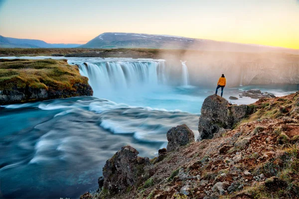 Adam gün batımında İzlanda 'da inanılmaz Godafoss şelalesinin yanında duruyor.